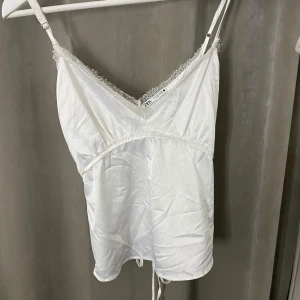 Vit satinlinnetopp från Zara med spets - Supersöt vit linnetopp i satin från Zara med tunna axelband och v-ringning dekorerad med spets. Toppen har justerbara band och snygg knytning i ryggen för extra detalj. Perfekt att styla med jeans eller kjol för en trendig look.
