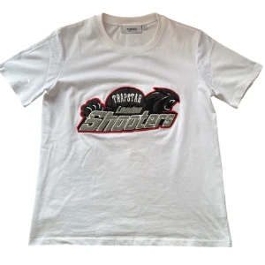 Vit t-shirt från Trapstar med broderad logga - Vit t-shirt från Trapstar med stort broderat motiv på bröstet där det står 'Trapstar London Shooters' och en svart panter. T-shirten har rund hals och korta ärmar med meshdetaljer på axlarna. Perfekt för en streetwear-look.