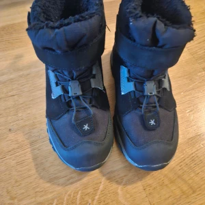 Svarta vinterkängor från Everest stl 30 - Svarta vinterkängor från Everest med varm fodring och praktisk snörning. Skorna har robust sula och förstärkt tå, perfekta för kalla dagar. Snygg och sportig design med detaljer i grått och svart. Storlek 30.