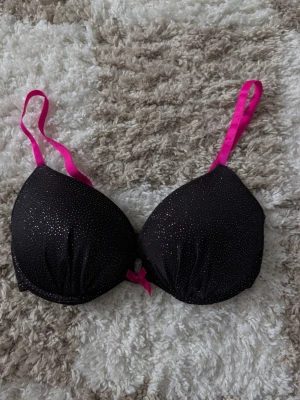 Svart glittrig bh med rosa detaljer - Snygg svart bh med glittriga prickar och starkt rosa axelband samt en liten rosett framtill. Bh:n har vadderade kupor och ger en cool och lekfull vibe. Perfekt för dig som gillar att sticka ut lite extra.