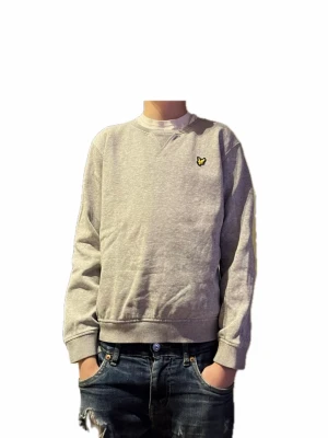 Grå sweatshirt från Lyle & Scott - Säljer en stilren grå sweatshirt från Lyle & Scott med klassisk rund hals och den ikoniska gula loggan broderad på bröstet. Tröjan har ribbade muddar vid ärmslut och nederkant, och är tillverkad i mjuk bomull för maximal komfort.