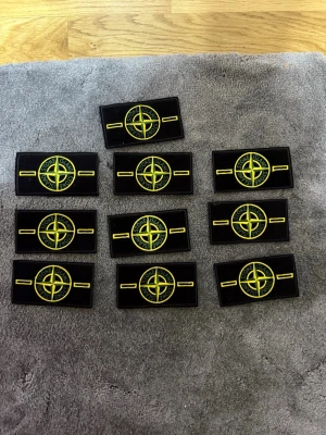 Stone Island patch till väst 120/kr styck - Säljer en klassisk svart Stone Island patch med gul och grön brodyr. Märket har den ikoniska kompassen och texten 'Stone Island' runt om. Perfekt att sy eller fästa på din väst för en streetwear-look.