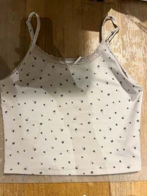 Ljusblommig linnetopp med spetskant - Supersöt ljus linnetopp med små mörkblå blommor och tunna axelband. Toppen har en fin spetskant upptill och en liten rosett i mitten. Perfekt till sommaren och enkel att matcha med jeans eller kjol.