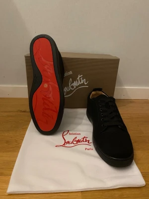 Christian Louboutin skor Helt Nya!!! - Säljer ett par svarta sneakers från Christian Louboutin med ikonisk röd sula. Skorna har snörning, rund tå och är tillverkade i mocka med detaljer i skinn. Insidan är beige och märkt med Louboutin-loggan. Kommer med originalkartong och dustbag.     Helt Nya!!!