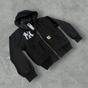 Carhartt x New York Yankees huvad jacka – Svart | M - Längd: 63-64 cm Bredd: 56 cm  Svart Carhartt-jacka med huva och stort New York Yankees-tryck på bröstet samt klassisk Carhartt-logopatch. Varm och slitstark med dragkedja, huva och ribbade muddar. Litet vit streck som syns på bilden! 