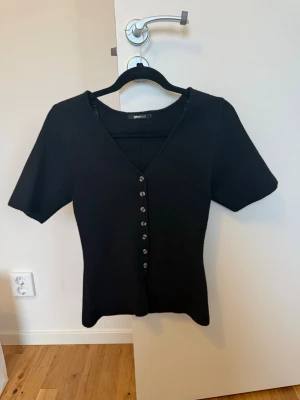 Svart topp med knappar Gina Tricot - Säljer en svart topp från Gina Tricot med v-ringning och korta ärmar. Toppen har en rad med knappar framtill och är figurnära i modellen. Perfekt att styla med jeans eller kjol för en clean look.