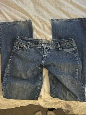 Lågmidjade bootcut jeans  - Säljer ett par blå bootcut jeans. Jeansen har ett hål på knät och slitningar längst ner på benen. Jeansen har klassisk femficksdesign, knappstängning och dekorativa bakfickor med knapp. 