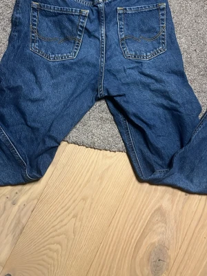 Blå jeans från Jack & Jones, relaxed fit - Säljer ett par klassiska blå jeans från Jack & Jones i relaxed fit. Jeansen har raka ben, fem fickor och kontrastsömmar. Tillverkade i slitstarkt denim med normal passform och midja. Perfekta för en chill och avslappnad stil.