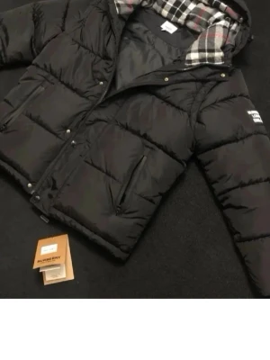 Svart dunväst från Burberry - Säljer en svart quiltad dunväst från Burberry med dragkedja och tryckknappar. Västen har två snedställda fickor med dragkedja och en avtagbar huva med klassiskt rutigt foder. Stor Burberry-patch på bröstet för extra statement.