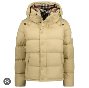 Burberry jacka  - Säljer en beige dunjacka från Burberry med klassiskt rutigt foder i huvan. Jackan har snörning vid huvan, dragkedja och knappar framtill samt två sidofickor med dragkedja. Puffad design och patch på ärmen ger en cool vibe.