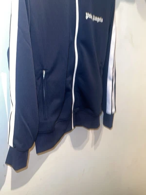 Palm Angels marinblå zip hoodie - Palm Angels marinblå hoodie med vita ränder längs ärmarna och vit dragkedja. Klassisk streetwear-look med logga på bröstet och två fickor framtill. Tillverkad i mjuk polyester som ger en sportig vibe.