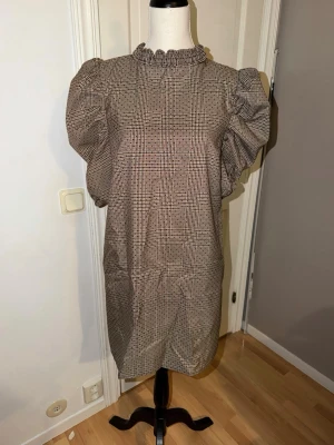 Rutig puffärmklänning från Zara - Säljer en rutig kort klänning från Zara med puffärmar och volang vid halsen. Klänningen har en beige, brun och svart färgskala och är tillverkad i ett vävt material. Baksidan har en snygg öppning med knappstängning. Perfekt för dig som gillar trendiga detaljer!
