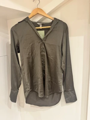 Mörkgrå skjorta i satinlook XS - Snygg mörkgrå skjorta från H&M i satinliknande polyester. Skjortan har klassisk krage, långa ärmar med manschetter och knappar framtill. Perfekt till jeans eller kjol för en clean och stilren vibe.