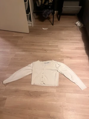Vit ribbad kofta från 157 - Säljer en ljusvit ribbad kofta från 157 med v-ringning och knappar framtill. Tröjan är croppad och har långa ärmar, perfekt att slänga över en topp eller linne. Enkel och clean stil som passar till det mesta.