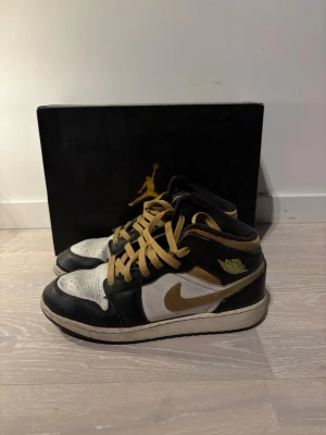 Nike Air Jordan 1 svart/vit/guld - Säljer ett par Nike Air Jordan 1 med svart, vit och guld färgskala. Skorna har klassisk hög siluett, guldfärgade snören och detaljer samt svart/vit ovandel i skinn. Ikonisk Jordan-logga på sidan och svart yttersula. Perfekta för dig som gillar streetwear och sneakerskultur.