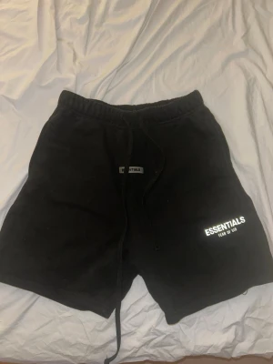 Svarta shorts Essentials Fear of God - Svarta shorts från Essentials Fear of God med tryckt logga på benet. De har elastisk midja med snörning och är gjorda i mjuk bomull. Perfekta för chill eller streetwear och har en clean, enkel look.
