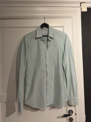 John Henric skjorta - Snygg skjorta från John Henric i grönvitt randigt mönster. Klassisk krage, långärmad och med knappar hela vägen framtill. Skjortan är i bomull och har en normal passform som sitter schysst på kroppen. Perfekt för dig som gillar stilrena och fräscha plagg.