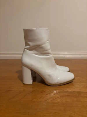 Vita boots med klack från NA-KD - Säljer ett par vita boots från NA-KD med fyrkantig tå och hög blockklack. Skorna är i syntetmaterial med glansig finish och har dragkedja på insidan. Perfekta för dig som vill sticka ut med en clean och trendig look.