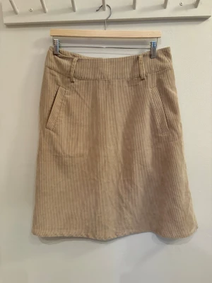 Beige manchesterkjol med fickor - Säljer en beige manchesterkjol med klassiska fickor fram och hällor för bälte. Kjolen har en rak, lätt A-linjeformad siluett och stängs med dragkedja bak. Perfekt för dig som gillar en enkel och stilren look.