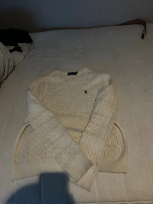 Vit kabelstickad tröja Polo Ralph Lauren - Kabelstickad vit tröja från Polo Ralph Lauren med klassisk rund halsringning och broderad logga på bröstet. Tröjan har långärmad passform och ribbade muddar. Perfekt för dig som gillar stilrena och tidlösa plagg.