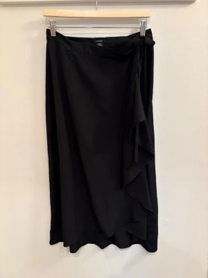 Svart omlottkjol från Lindex - En svart långkjol från Lindex i omlottmodell med knyt i midjan. Kjolen har en stilren och enkel design med lätt fallande tyg och en diskret volangdetalj framtill. Perfekt för dig som gillar minimalistisk stil och vill ha något som känns både bekvämt och snyggt.