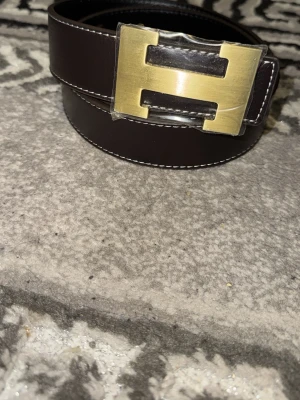 Brunt bälte med guldfärgat H-spänne - Sny soprent brunt bälte i skinn med ett stort guldfärgat H-spänne. Bältet har vita sömmar längs kanterna som ger en clean kontrast. Perfekt accessoar för att lyfta vilken outfit som helst.