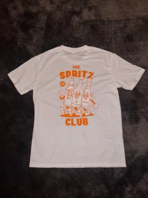 The Spritz Club t-shirt - t-shirt med trycket 'The Spritz Club' i orange fram och stort motiv på ryggen med tecknade flaskor och text. Klassisk passform och rund hals. Perfekt för dig som gillar coola prints och vill sticka ut lite extra.