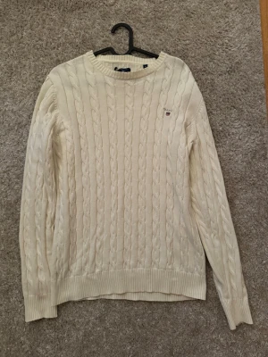 Vit kabelstickad tröja från Gant - Kabelstickad vit tröja från Gant med rund halsringning och diskret logga broderad på bröstet. Tröjan har långa ärmar och ribbade muddar vid ärmslut och nederkant. Perfekt för lager-på-lager och klassisk stil.