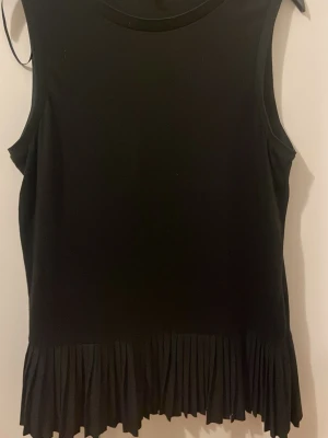 Svart ärmlös topp från STOCKH LM - Säljer en svart ärmlös topp från STOCKH LM i storlek M. Toppen har en rund halsringning och en snygg plisserad volangkant nedtill som ger en extra detalj. Perfekt att styla med jeans eller kjol för en clean look.
