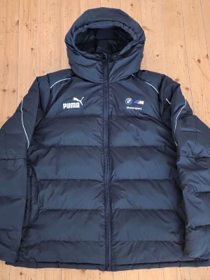 Svart BMW Motorsport pufferjacka Puma - Säljer en svart pufferjacka från Puma i samarbete med BMW Motorsport. Jackan har huva, dragkedja framtill och vita detaljer samt loggor på bröstet. Tillverkad i polyester och perfekt för kalla dagar. Snygg och sportig look för dig som gillar motorsport.
