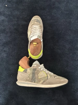 Camo sneakers från Philippe Model - Säljer ett par Philippe Model sneakers i beige och grönt camomönster med detaljer i mocka och neon-gult vid hälen. Skorna har snörning, rund tå och platt sula. Klassisk logga på sidan och cool urban vibe. Kommer med box och knappast använda