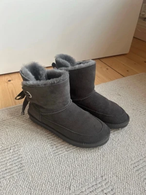 Mörkgrå boots från Gooce med rosett - Säljer ett par mörkgrå boots från Gooce med fluffigt foder och snygga rosetter bak. Skorna är i mocka och har en rejäl, mönstrad sula för bra grepp. Perfekta för kalla dagar och riktigt mysiga att ha på sig.