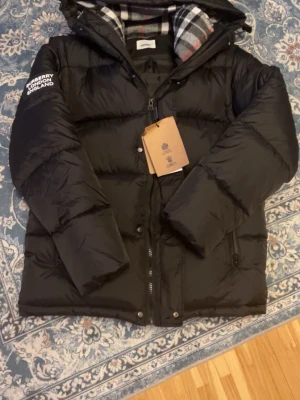 Svart pufferjacka från Burberry - Säljer en svart pufferjacka från Burberry med klassiskt rutigt foder i huvan. Jackan har dragkedja framtill, huva och logga på ärmen. Perfekt för kalla dagar och riktigt snygg till streetwear-looken.