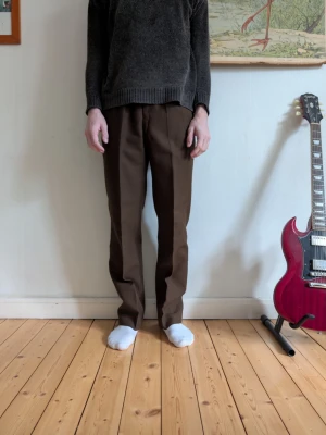 Bruna kostymbyxor med pressveck - Säljer ett par bruna kostymbyxor med klassiskt rakt snitt och pressveck framtill. Byxorna har en enkel, stilren look och är tillverkade i ett mjukt tyg som känns bekvämt. Perfekta för dig som vill ha en clean och tidlös stil.