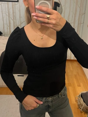 Svart ribbad topp från H&M - Säljer en svart långärmad topp från H&M i storlek XS. Toppen är ribbad med rundad, djup urringning och har en tight passform & små puffärmar. Perfekt att styla med jeans eller kjol för en clean look.