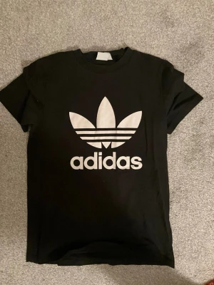 Svart Adidas t-shirt med logga - Säljer en svart t-shirt från Adidas med den klassiska vita trefoil-loggan och texten på bröstet. T-shirten har rund hals och korta ärmar, perfekt för en sportig och avslappnad stil. Tillverkad i mjuk bomull för skön känsla. Köpt för 300 säljs för 80 kr!