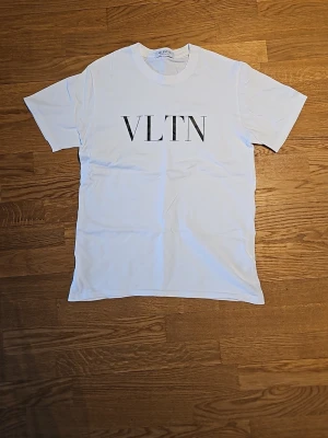 Vit VLTN t-shirt från Valentino - Snygg vit t-shirt från Valentino med stor svart VLTN-print på bröstet. Klassisk rak passform och korta ärmar. Tillverkad i mjuk bomull och made in Italy. Perfekt för dig som gillar stilrena designer och exklusiva märken.