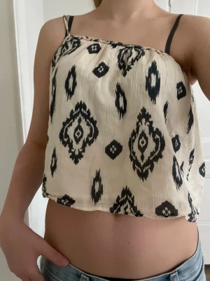 Beige croppad linnetopp med mönster - Säljer en beige croppad linnetopp med svart grafiskt mönster. Toppen har tunna axelband och är tillverkad i ett lätt, luftigt material som passar perfekt till sommaren. Den har en loose passform och är ärmlös för en avslappnad vibe.