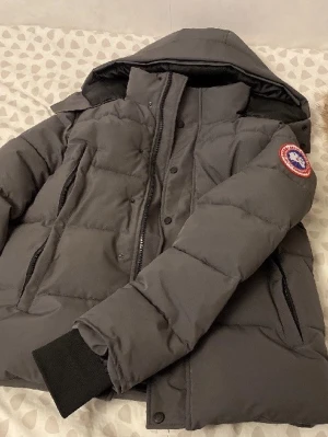 Mörkgrå Canada Goose dunjacka - Säljer en mörkgrå dunjacka från Canada Goose med avtagbar pälsdetalj till huvan. Jackan har klassisk patch på ärmen, dragkedja och knappar framtill samt ribbade muddar vid ärmsluten. Perfekt för kalla vinterdagar och riktigt snygg passform.