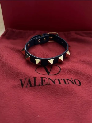 Valentino mörkblå armband - Säljer ett mörkblått armband från Valentino med ikoniska guldfärgade pyramidnitar. Armbandet är gjort i läder och har justerbart spänne. Perfekt accessoar för att ge din outfit en edgy touch.