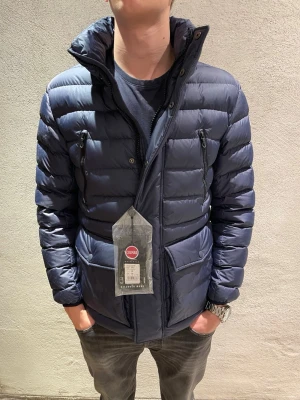 Mörkblå pufferjacka från Colmar - Säljer en mörkblå pufferjacka från Colmar med hög krage och dragkedja framtill. Jackan har två bröstfickor med dragkedja och två sidofickor med knappar. Snygg, quiltad design och perfekt för kyliga dagar.