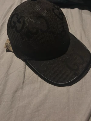 Svart Gucci keps med GG-mönster - Säljer en svart keps från Gucci med ikoniskt GG-mönster över hela kepsen. Justerbar rem bak i skinn och detaljer i svart. Tillverkad i slitstarkt material och riktigt snygg till streetwear-looken. Den är äkta 
