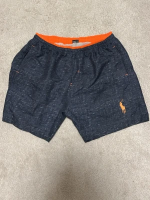 Badshorts från Polo Ralph Lauren 3XL - Mörkblå badshorts från Polo Ralph Lauren med orange detaljer och broderad logga.  De är xl men passar L .Perfekta för stranden eller poolen, riktigt snygg färgkombo och klassisk design. Helt oanvända nyskick