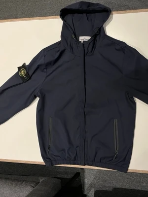 Mörkblå vindjacka från Stone Island - Säljer en mörkblå vindjacka från Stone Island med huva och ikonisk patch på vänster ärm. Jackan har dragkedja framtill, två fickor med dragkedja och elastisk nederkant. Tillverkad i polyester och har en clean, stilren look som passar perfekt till streetwear.