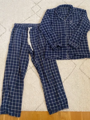 Blåvit rutig pyjamas från Change - Säljer en klassisk blåvit rutig pyjamas från märket Change. Skjortan har krage, bröstficka med broderad logga och knappar framtill. Byxorna har vida ben, resår i midjan och vit dragsko för skön passform. 
