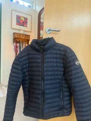Mörkblå pufferjacka från Colmar - Säljer en mörkblå pufferjacka från Colmar Originals. Jackan har quiltad design, hög krage och två sidofickor med dragkedja. Klassisk logga på ärmen och hel dragkedja framtill. Perfekt för kyliga dagar och riktigt snygg till alla outfits.