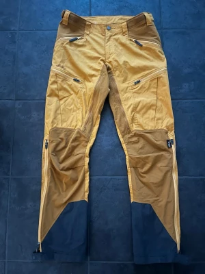 Lundhags jaktbyxor  - Säljer ett par gold dark/ gold Lundhags byxor med flera praktiska fickor, dragkedja, justerbar midja och är perfekta för utomhusaktiviteter. Materialet är slitstarkt och passar för tuffare väder. Använda väldigt lite så är som nya. 