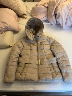Beige jacka Hollies Mont Blanc stl 36 - Säljer en beige pufferjacka från Hollies modell Mont Blanc med guldfärgade dragkedjor och bälte i midjan. Jackan har två bröstfickor och två sidofickor med dragkedja. Det finns även en pälskrage som skickas med. Bältet är uttöjt så det sitter inte så bra på tyvärr. Även lite fläckar på framsidan som kanske eventuellt går bort vid kemtvätt. Annars är jackan i fint skick. Storlek 36.
