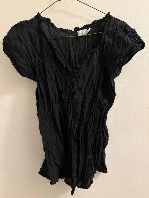 Svart blus med knappar och volang - Säljer en svart blus med korta puffärmar och v-ringning. Blusen har dekorativa volangdetaljer längs halsringningen och knappar framtill. Tyget är lätt och har en skrynklig struktur som ger en avslappnad känsla.