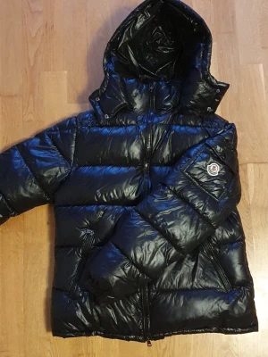 Svart Moncler pufferjacka med huva - Säljer en svart pufferjacka från Moncler med glansig finish och stor huva. Jackan har dragkedja framtill, två snedställda fickor med dragkedja och en Moncler-logga på ärmen. Perfekt för kalla dagar och riktigt snygg till streetwear.